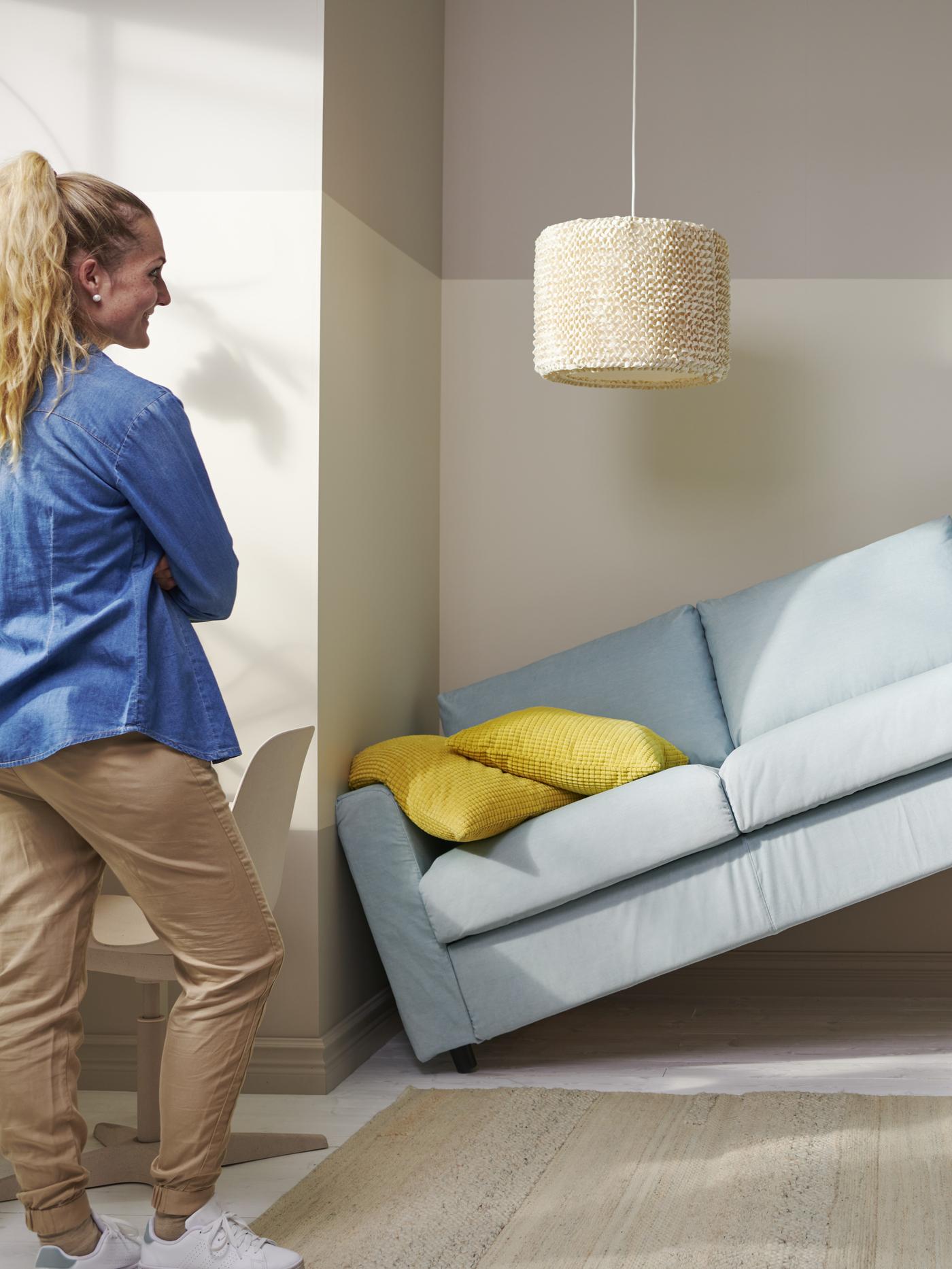 Kobieta stoi naprzeciwko błękitnej sofy, która jest przechylona pod kątem, opierając się o ścianę i kolumnę w nowoczesnym, jasnym wnętrzu z żółtą poduszką na sofie i plecionym pufem na podłodze.