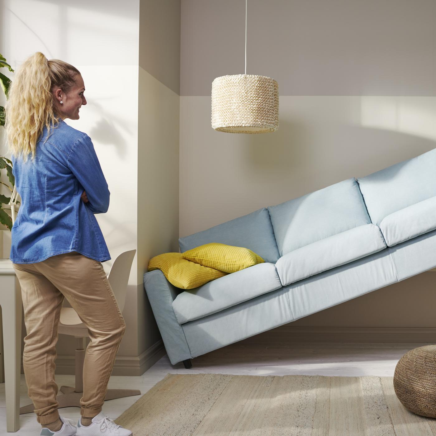 Kobieta stoi naprzeciwko błękitnej sofy, która jest przechylona pod kątem, opierając się o ścianę i kolumnę w nowoczesnym, jasnym wnętrzu z żółtą poduszką na sofie i plecionym pufem na podłodze.