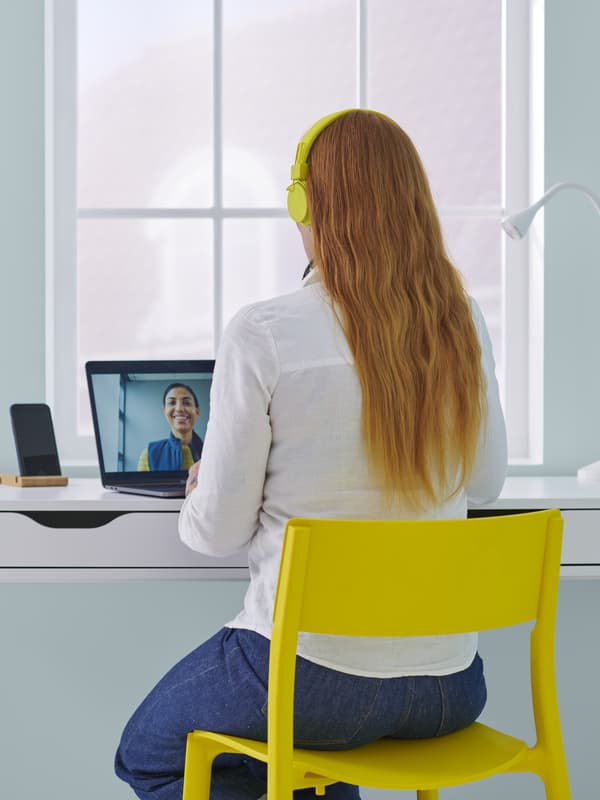 Kobieta w białej bluzce i długich włosach siedzi skierowana w stronę okna na żółtym krześle przy biurku i rozmawia z kimś na spotkaniu online na laptopie.