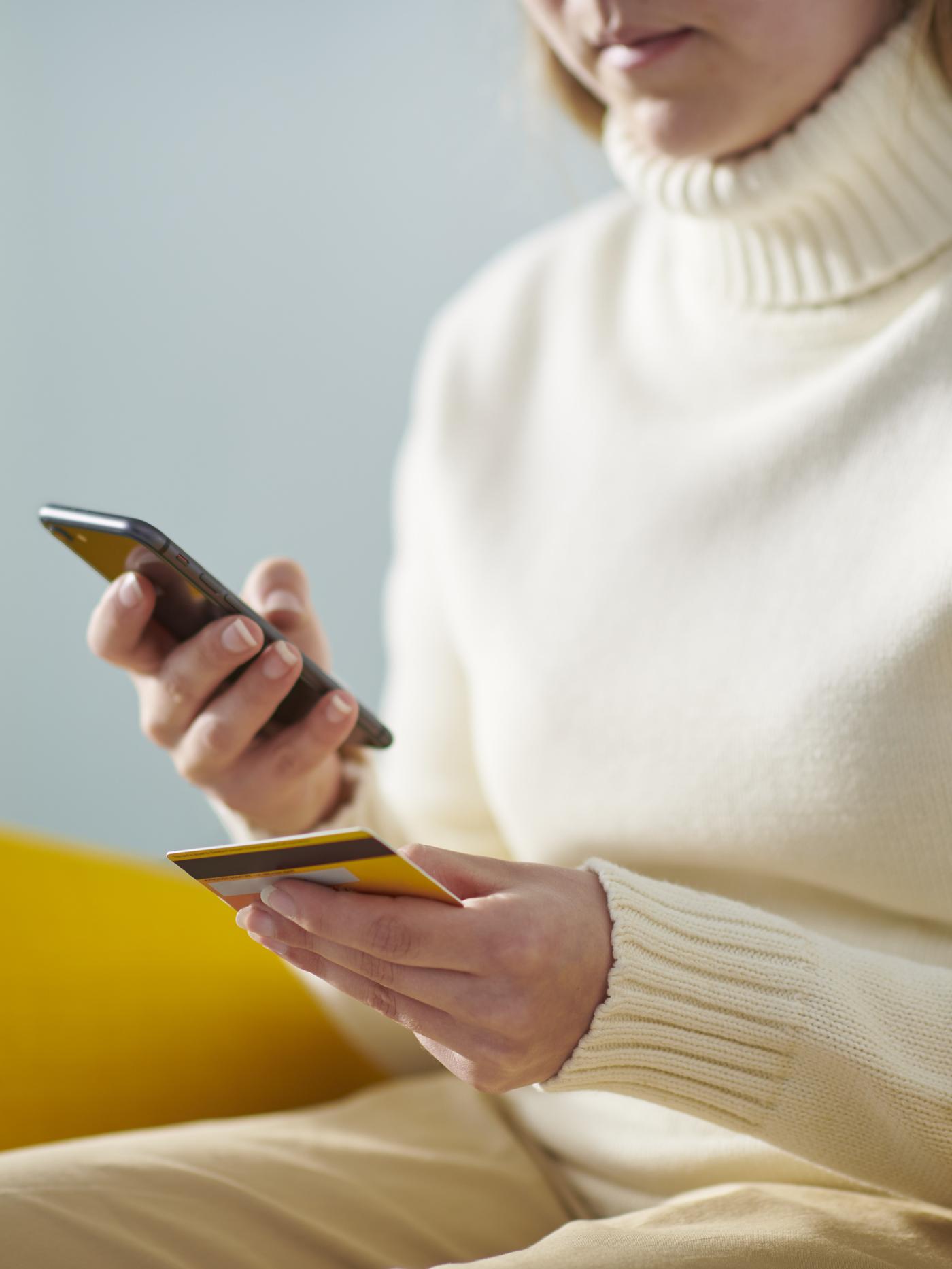 Kobieta w kremowym golfie w prawej ręce trzyma telefon, a w lewej kartę płatniczą.