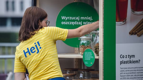 Kobieta w żółtej koszulce IKEA z napisem "Hej!" na plecach rozkłada towar w sklepie IKEA