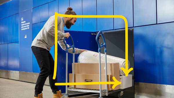 Kolica za transport s metalnim ručkama na kojima se nalazi velika kartonska kutija s IKEA logotipom i zamotani madrac, u pozadini plavi zid s ormarićima i natpisom.