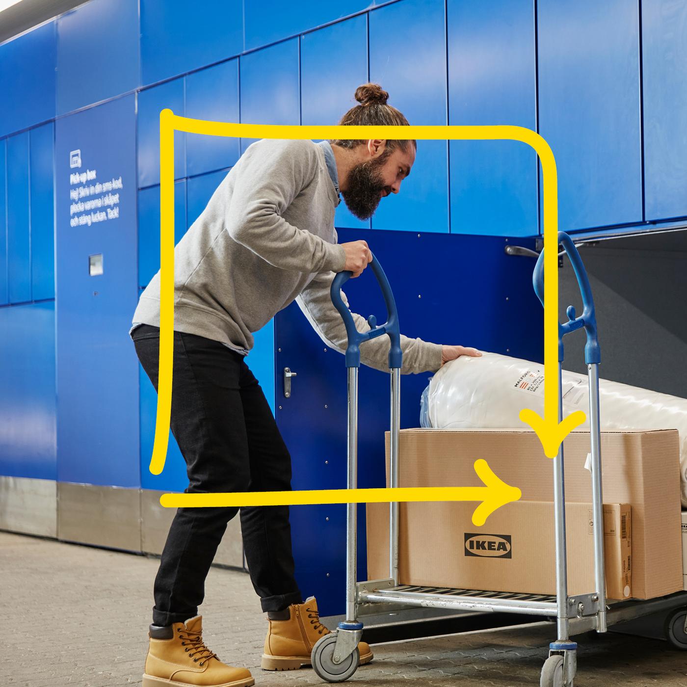 Kolica za transport s metalnim ručkama na kojima se nalazi velika kartonska kutija s IKEA logotipom i zamotani madrac, u pozadini plavi zid s ormarićima i natpisom.