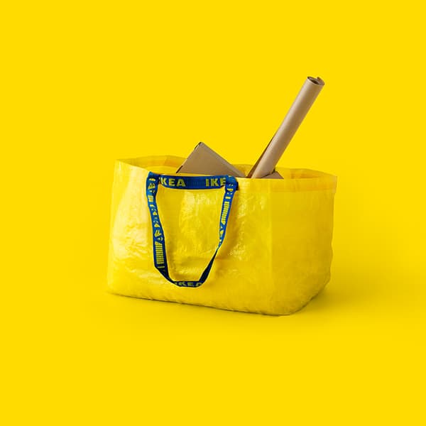 Kollane IKEA ostukott siniste käepidemetega, täidetud papist pakendi ja rullitud pabertoruga, asetatud ühtlasele kollasele taustale.