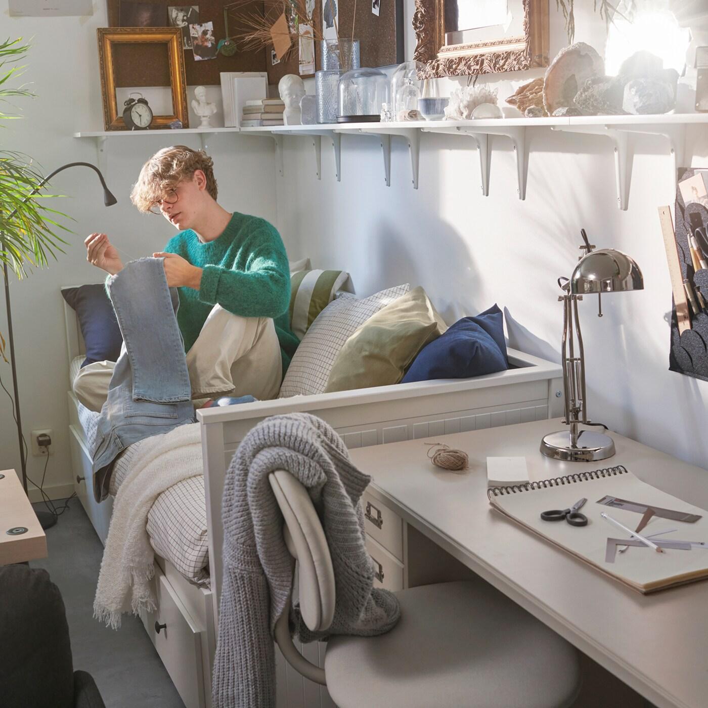 Kollégiumi szobában HEMNES kanapéágyon ülő diákot látunk. Felette több BURHULT/SIBBHULT polc kapott helyet, rajtuk képkeretek, könyvek és dekorációk.