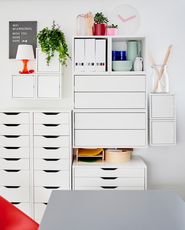 Kombinácia bielych nástenných skriniek a bielych zásuvkových dielov so stolovacím riadom, dekoráciami a zakladačmi na časopisy. 