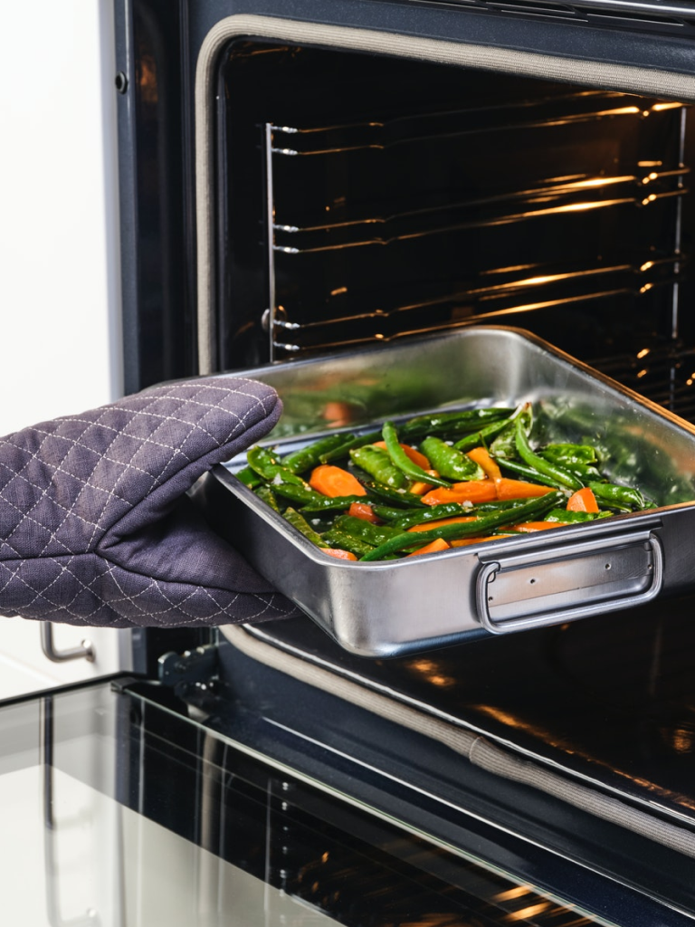 오븐 장갑을 낀 한 손이 당근과 다른 채소가 채워진 KONCIS 콘시스 로스팅팬을 오븐에 넣고 있는 모습.