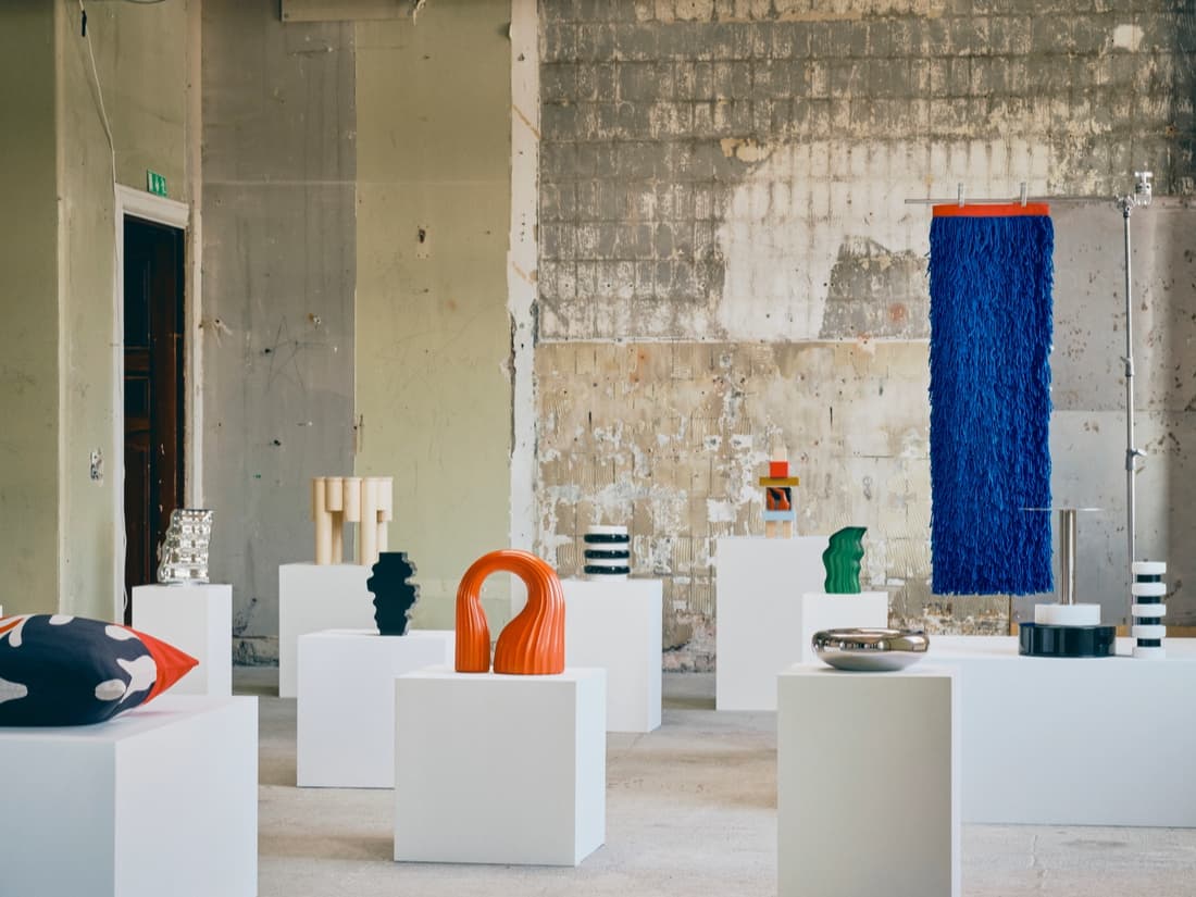 KONSTRUNDA art exhibition with various abstract sculptures and objects displayed on white pedestals in an industrial space with a textured, unfinished wall.
