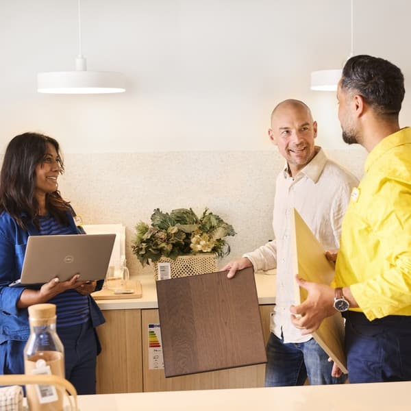 Konsultant IKEA w żółtej koszulce i pracowniczka IKEA w niebieskiej koszuli prowadzący usługę planowania kuchni w domu klienta. Obok konsultanta stoi klient trzymający front kuchenny w ręku.