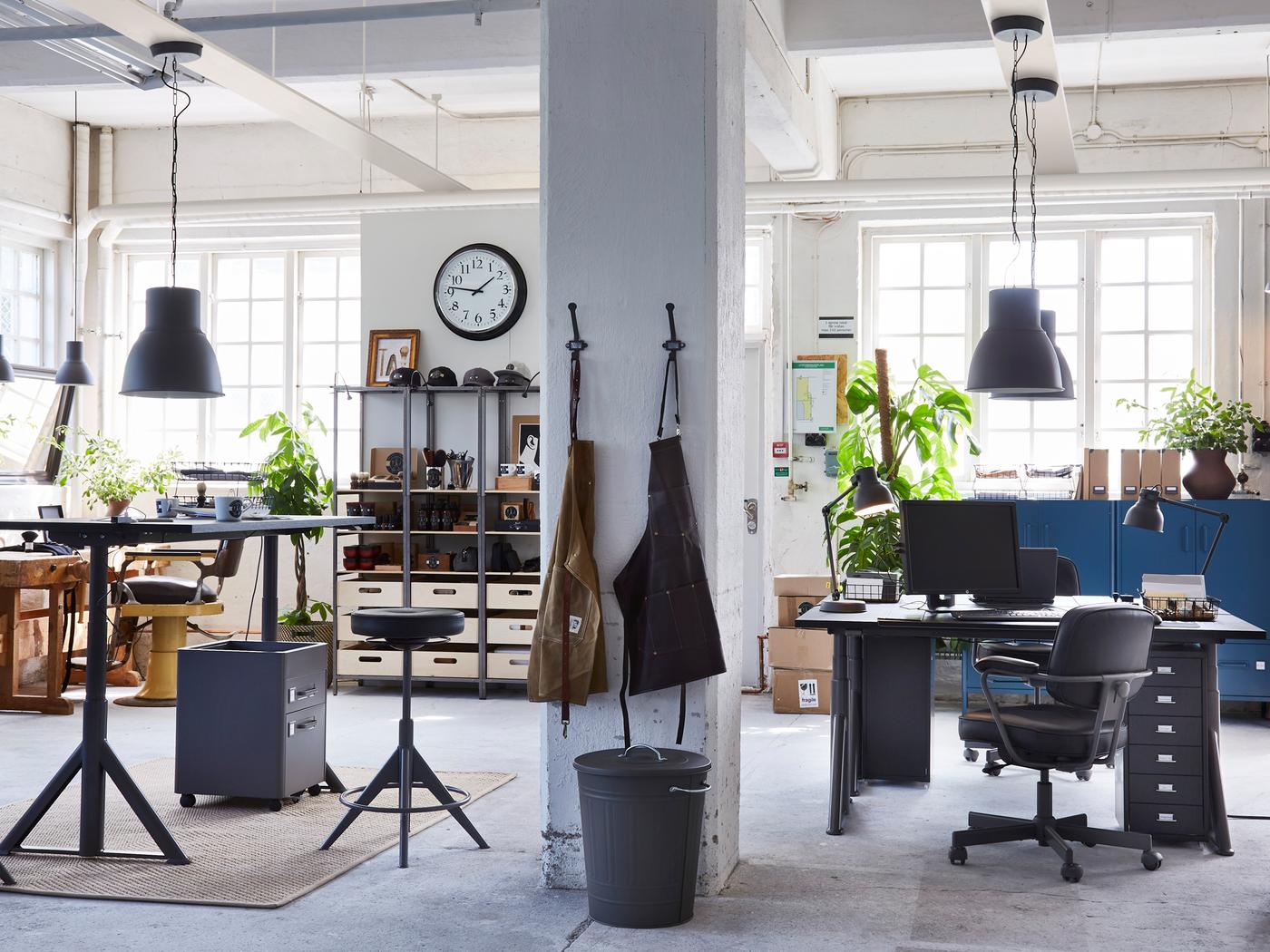 Kontor i industriell stil med hyller, arbeidsplasser med IDÅSEN skrivebord sitte/stå og forklær som henger på ei betongsøyle. 