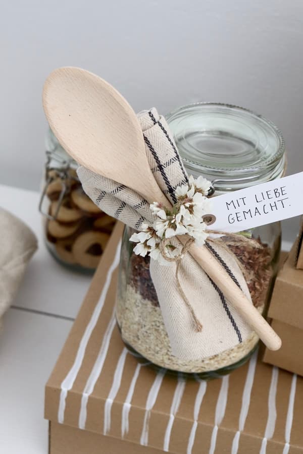 KORKEN Vorratsglas befüllt mit einer Backmischung. Daran befestiht mit einem Geschenkanhänger und Schnur: EIn Kochlöffel aus Holz, ein Geschirrtuch zusammengerollt uns ein kleines Sträußchen Trockenblumen.