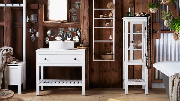 Koupelna s tmavými dřevěnými stěnami, pokrytá malými zrcadly různých tvarů a bílým koupelnovým nábytkem HEMNES.