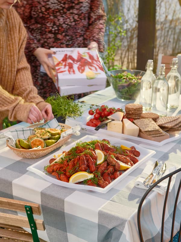 โต๊ะที่มีแสงแดดส่องจัดเตรียมไว้สำหรับงานเลี้ยง มีกุ้งเครย์ฟิช KRÄFTOR วางอยู่บนถาด SMULA/สมูล่า ชีสและแคร็กเกอร์อยู่บนถาดอีกใบ