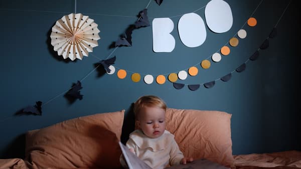 Kreativt pyssel till halloween av papper, girlanger, vimplar och pappers diy hänger på väggen ovanför en säng. I sängen sitter ett litet barn och läser en bok.