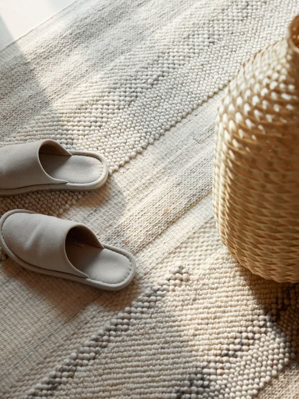 Krémszínű papucsok és egy szőtt bambusz váza áll a kézzel szőtt BRÖNDEN szőnyegen.