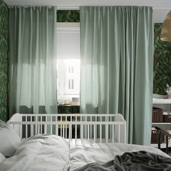 Krevet i krevetac se nalaze pored prozora sa zelenim/belim zavesama i belim roletnama za zamračivanje koje pokrivaju pola prozora.