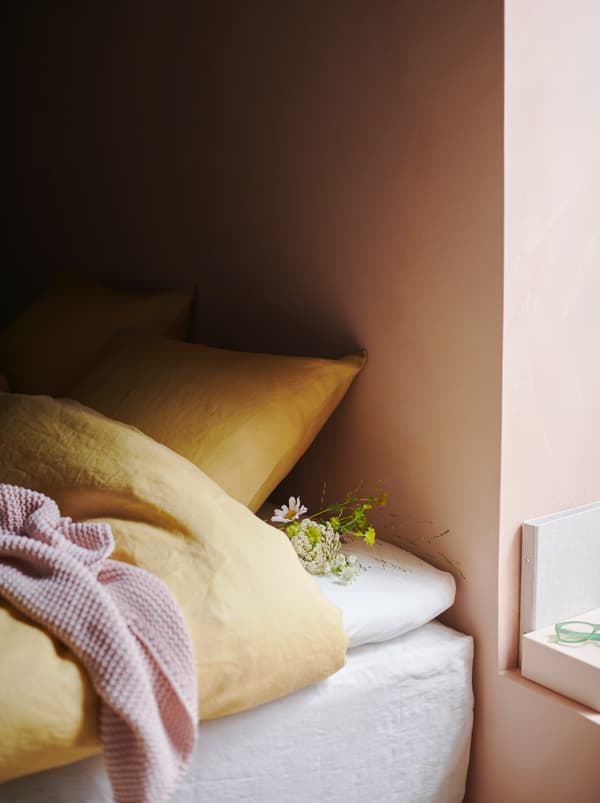 Krevet na kojem se nalazi žuta PUDERVIVA posteljina, roza INGABRITTA lagana deka na njoj te mali buket cvijeća koji leži pokraj jastuka.