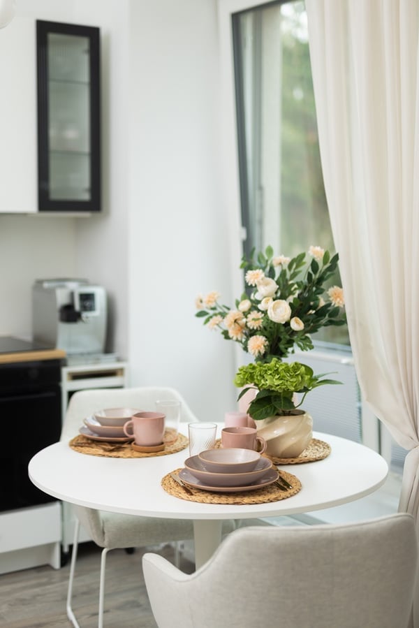 Круглый стол у окна, на столе — посуда и ваза с букетом цветов; занавески на окне добавляют уют.