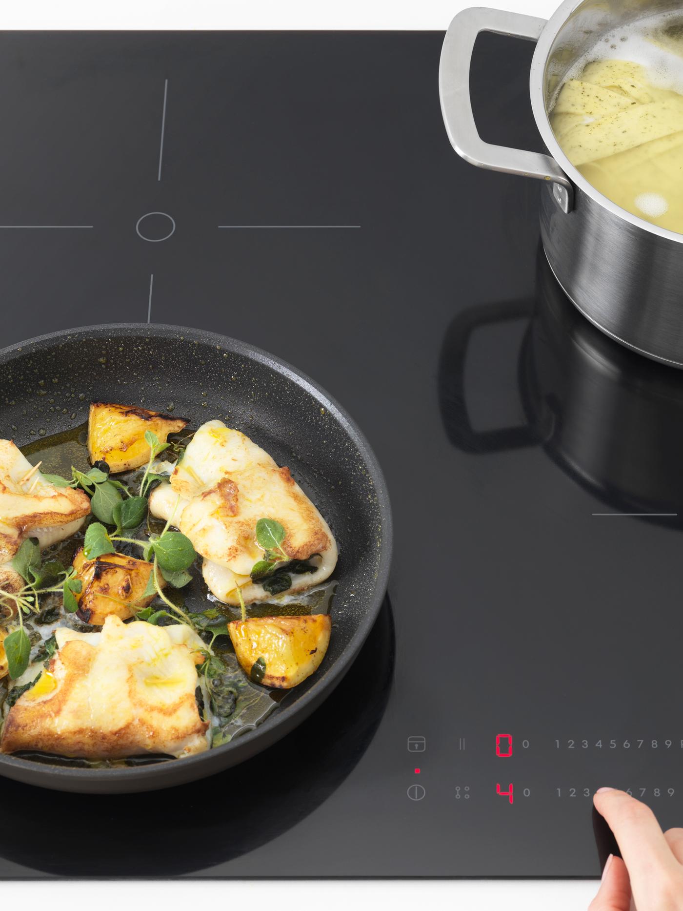Krupan kadar indukcijske ploče s crnim tiganjem u kojem se prži riba i srebrnom posudom s testeninom.