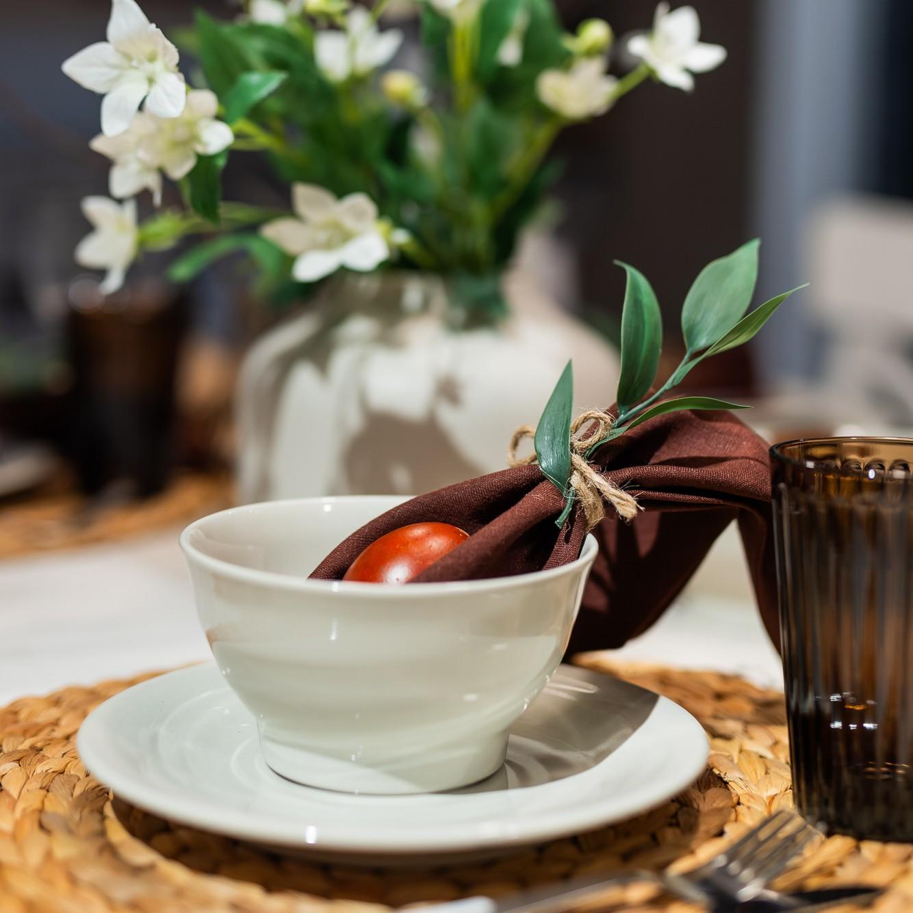 Крупный план белой тарелки на плетёной подставке, сверху — белая миска, окрашенное яйцо и салфетка с зелёной веточкой внутри.