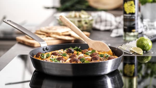 Kuali leper dengan pemegang keluli tahan karat berisi bebola daging bersos yang sedang dimasak di atas hob. Sudu kayu terletak di sisi.
