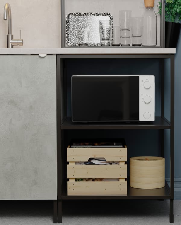 Kuchenka mikrofalowa, drewniana skrzynka i drewniane pudełko ustawione na otwartych półkach pod blatem kuchennym. Obok widoczna jest szafka z drzwiami imitującymi beton.
