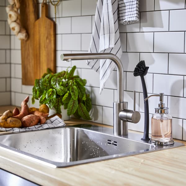 Kuchynská batéria vo farbe nehrdzavejúcej ocele, drez z nehrdzavejúcej ocele, kefka na umývanie riadu a čierno-biele kuchynské utierky.