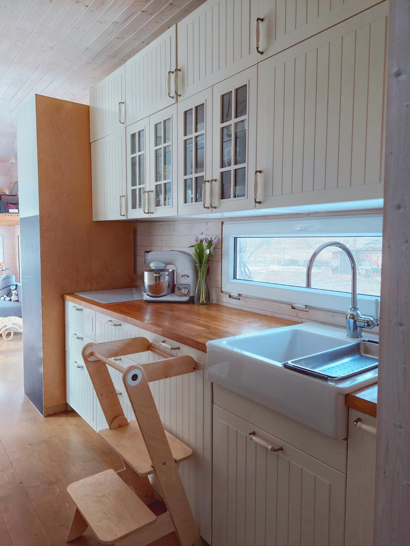 Kuchyňská linka v béžové barvě s velkým, porcelánovým dřezem, s deskou v dřevěném dekoru a nástěnnými skříňkami až po strop. K lince jsou postaveny schůdky ze dřeva, aby se snáž dosáhlo na vrchní úložné prostory.