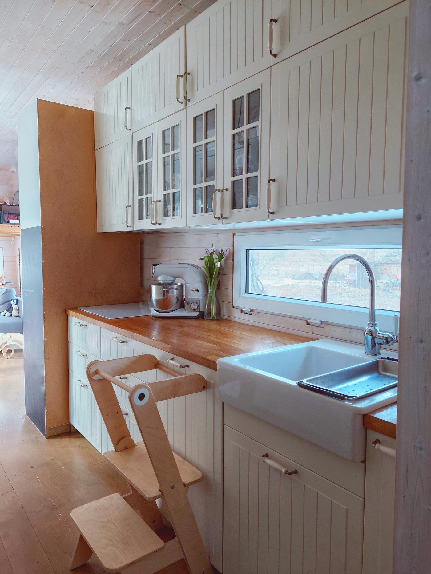 Kuchynská linka v béžovej farbe s veľkým, porcelánovým drezom, s doskou v drevenom dekore a nástennými skrinkami až po strop. K linke sú postavené schodíky z dreva, aby sa ľahšie dosiahlo na vrchné úložné priestory.