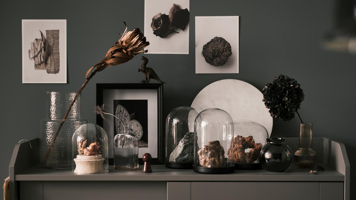 Különböző kövek SKÖNJA üvegbúrák alatt, egy szürke HAUGA üvegajtós szekrény tetején, mellettük üvegvázákban virágok.