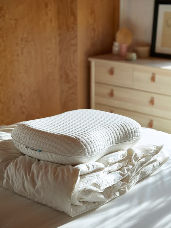 Kuilt dan bantal KLUBBSPORE ditindan bersama di atas duvet bersih berwarna putih di dalam bilik yang disinari cahaya matahari.