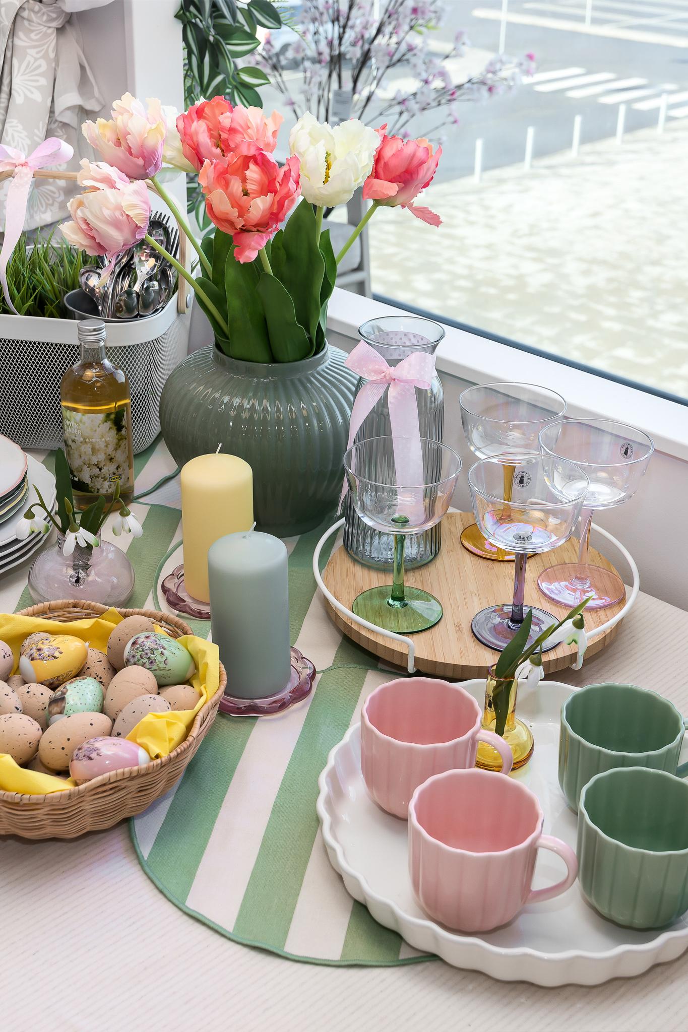 Кухонный стол, украшенный к Пасхе: ваза с тюльпанами, пастельные кружки, свечи, стеклянная посуда и корзина с разноцветными декоративными яйцами.