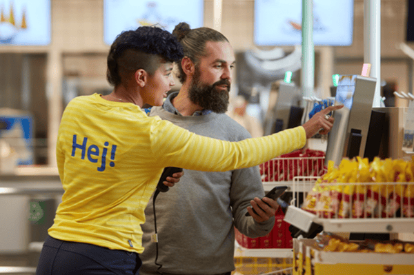 Kund får hjälp i kassan av en IKEA medarbetare med en arbetströja med ordet Hej på ryggen på sig.