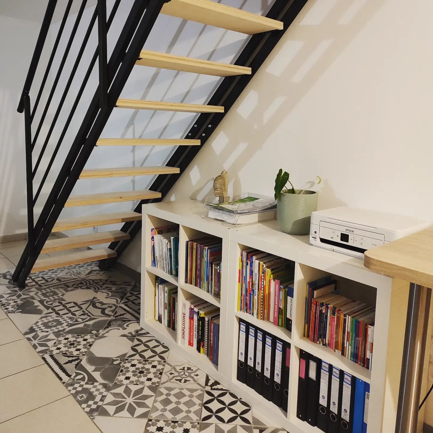 Kundbild av en vit bokhylla under trappa med böcker och pärmar, dekorerad med växt och små föremål. Svart metalltrappa med trästeg och mönstrat golv ger en modern och praktisk känsla.
