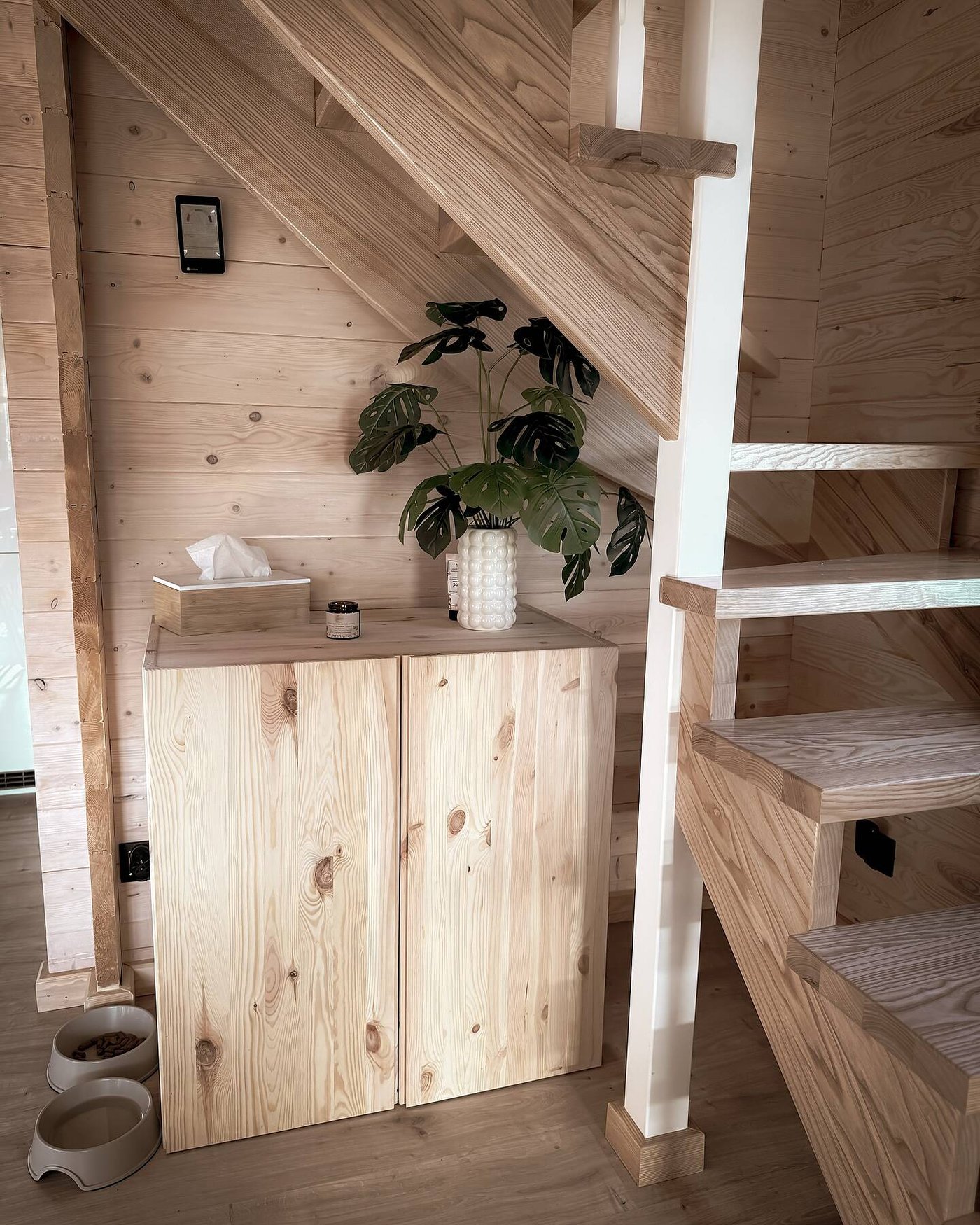 Kundbild av ljus träförvaringsmöbel under trappa i matchande träpanel. Dekorerad med grön växt i vit kruka, servettlåda och ljus, med matskålar på golvet för husdjur.
