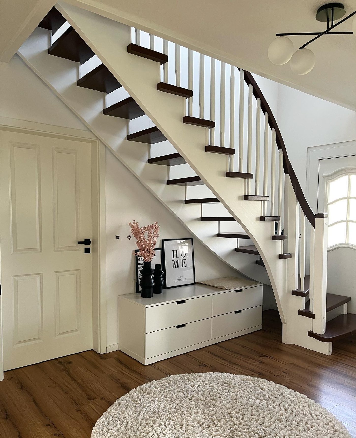 Kundbild av vit förvaringsmöbel med lådor under svängd trappa i trä och vitt räcke. Dekorerad med svart vas, torkade blommor och tavla, på golv i trä med rund matta.
