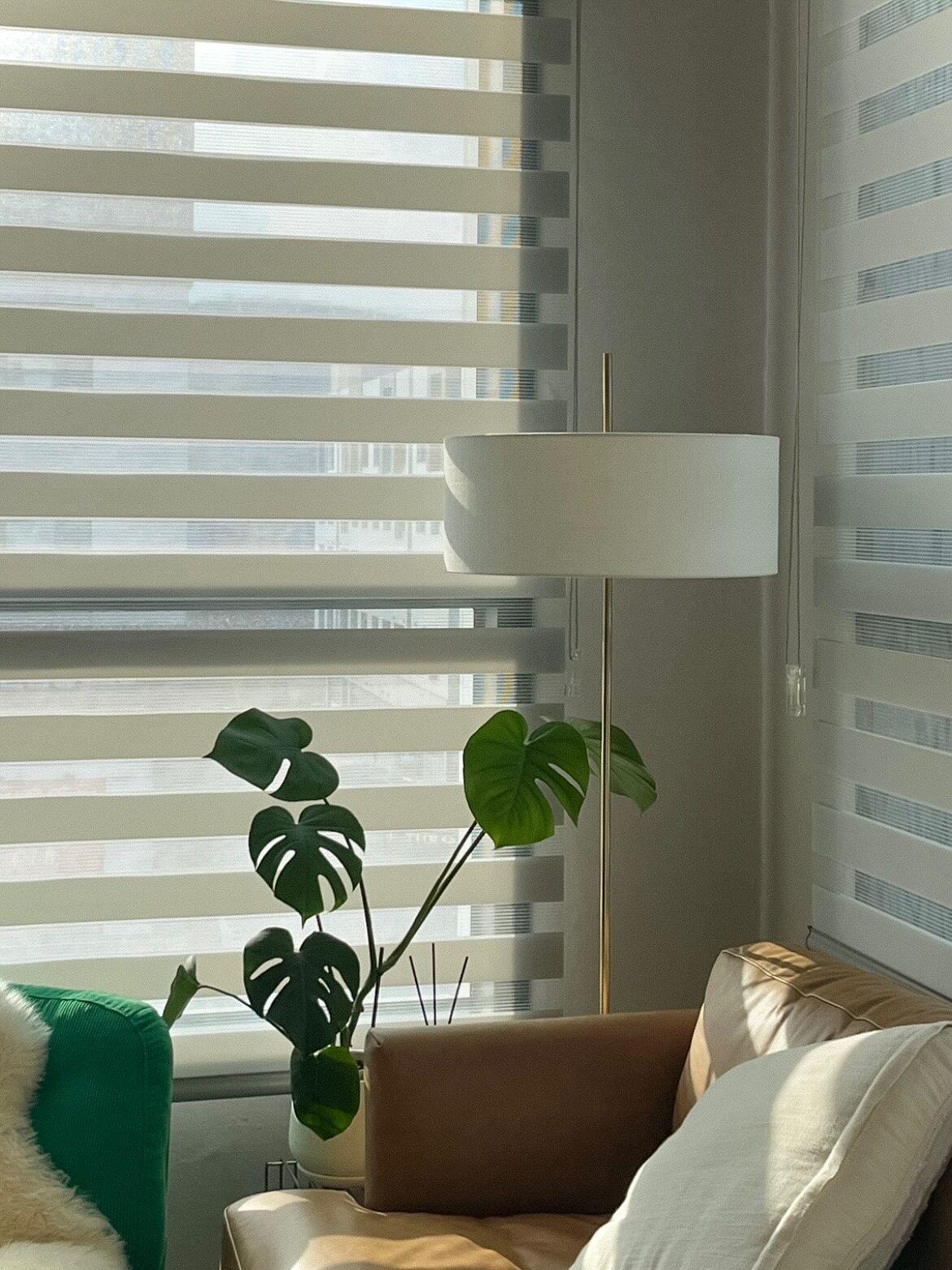 Kundbild från @patrycja.brylka på en vardagsrumshörna med en brun lädersoffa, en vit STOCKHOLM 2025 golvlampa och en monsteraplanta vid ett fönster med randiga persienner.