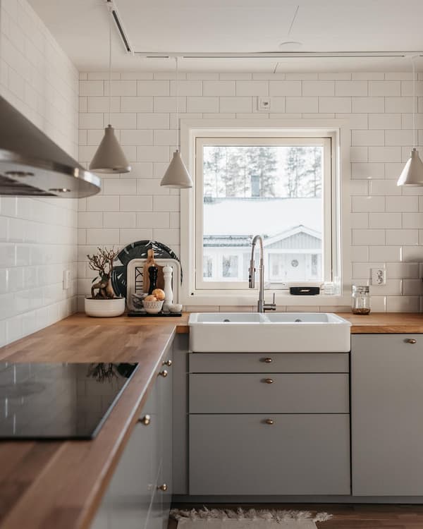 Kundbild på kök med lantlig diskho, träbänkar, grå skåp, vitt kakel och utsikt mot snötäckta träd genom fönstret.