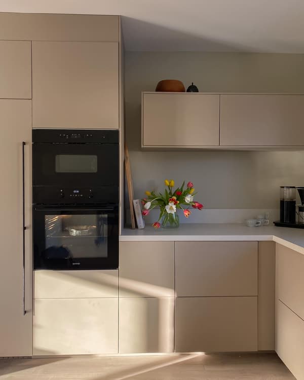 Kundbild på modernt kök med beige skåp, svart inbyggd ugn, tulpanvas, kaffebryggare och solskuggor på golvet.