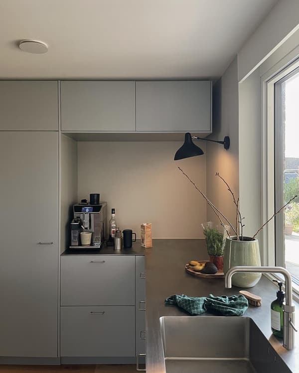 Kundbild på modernt kök med grå skåp, kaffemaskin, diskho med grön trasa, växter i vaser och naturligt ljus från fönster.