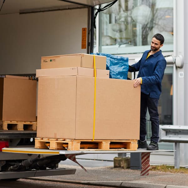 Kuorma-auton lavalla seisoo mies, joka toimittamassa isoa postipakettia asiakkaalle.