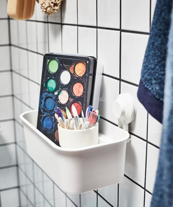 Kutija vodenih boja i šalica puna kistova u košari s vakuumskim držačima na kupaonskom zidu s pločicama uz potrepštine za tuširanje.