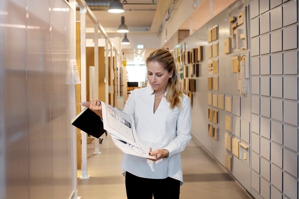 Kvinna i vit skjorta står i en korridor med ett svart anteckningblock och olika ritningar