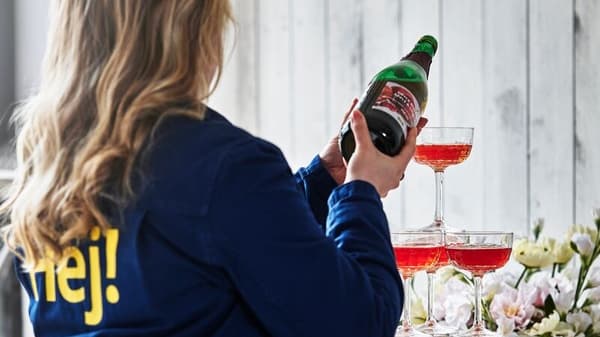 Kvinna med blå IKEA-jacka med texten Hej på ryggen häller upp röd tryck i ett torn av glas.
