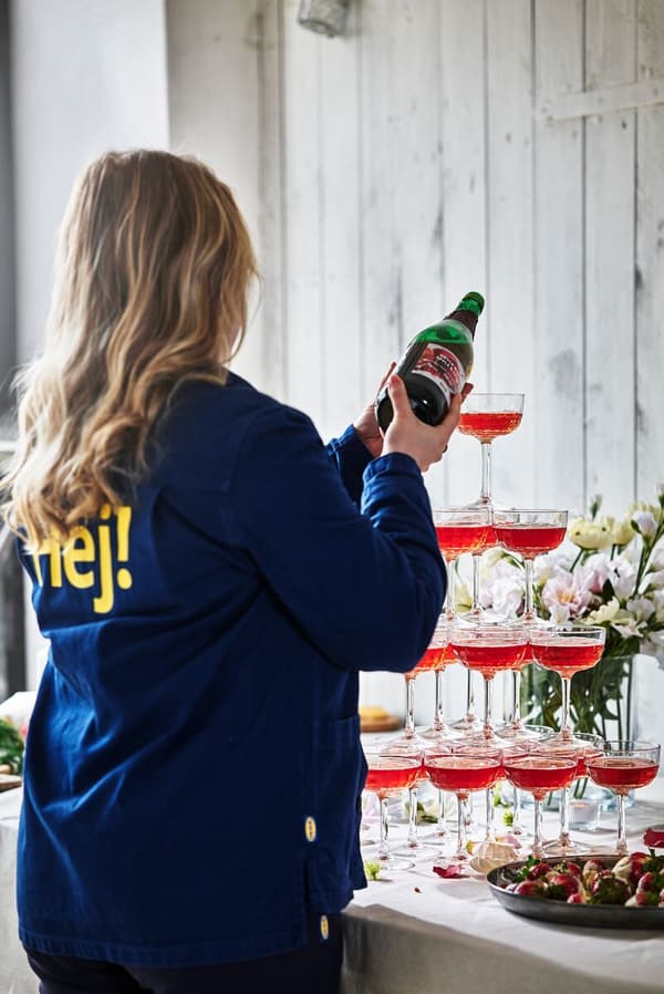 Kvinna med blå IKEA-jacka med texten Hej på ryggen häller upp röd tryck i ett torn av glas.