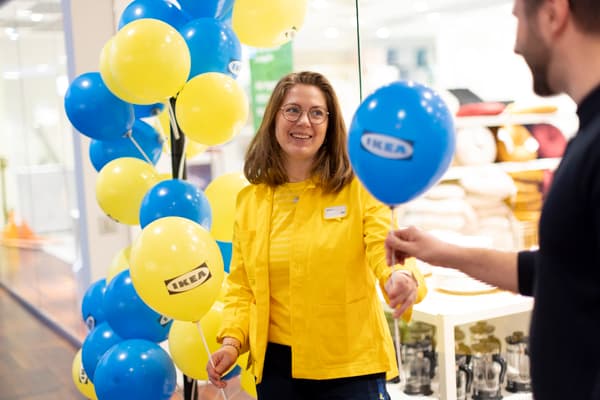 Kvinna med gula kläder står framför en grupp ballonger och delar ut en blå ballong till en kund