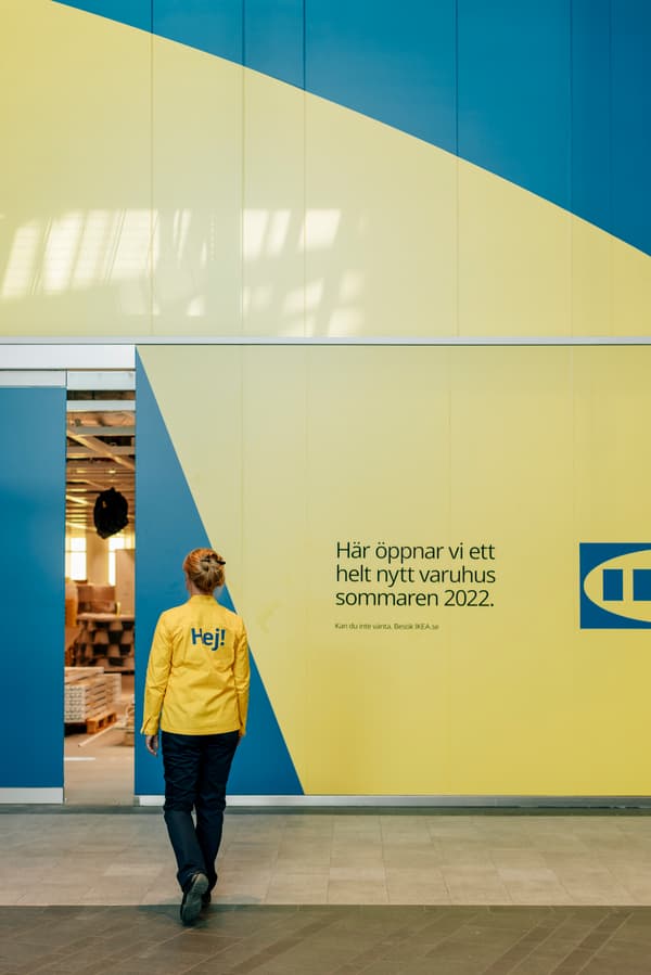 Kvinna med  texten "Hej!" på ryggen tittar på en stor gul vägg som har en liten öppen glipa