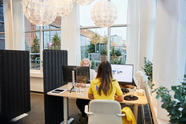 Kvinna sitter med gul tröja med ryggen vänd och tittar mot en skärm. På andra sidan bordet sitter en kvinna vid en skärm.
