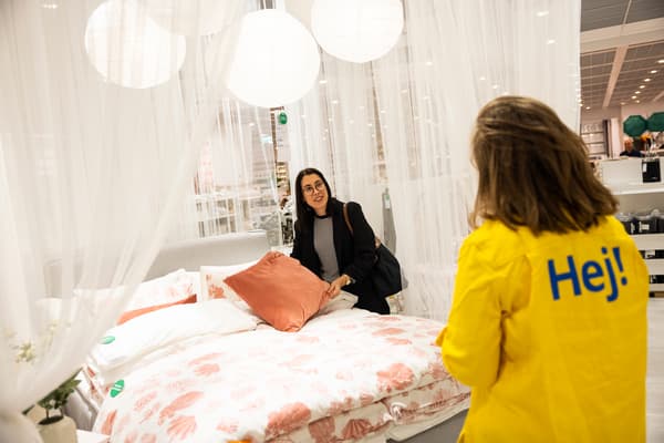 Kvinna står vid säng och tittar på en orange kudde, medan en medarbetare med gula kläder och texten "hej!" på ryggen står och pratar med henne