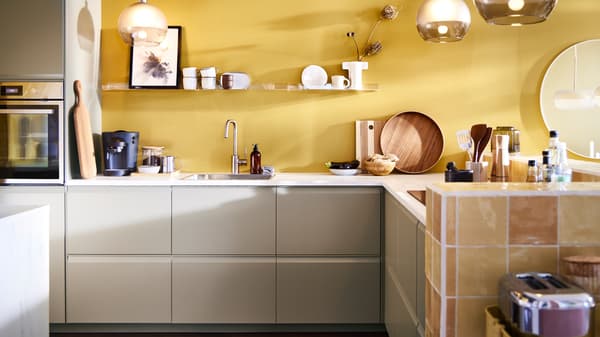 L-Form Küche mit UPPLÖV Schubladenfronten in mattem Dunkelbeige, Hängeleuchten mit JAKOBSBYN Hängeleuchtenschirmen in Frostglas und Grau und andere Küchengegenständen.